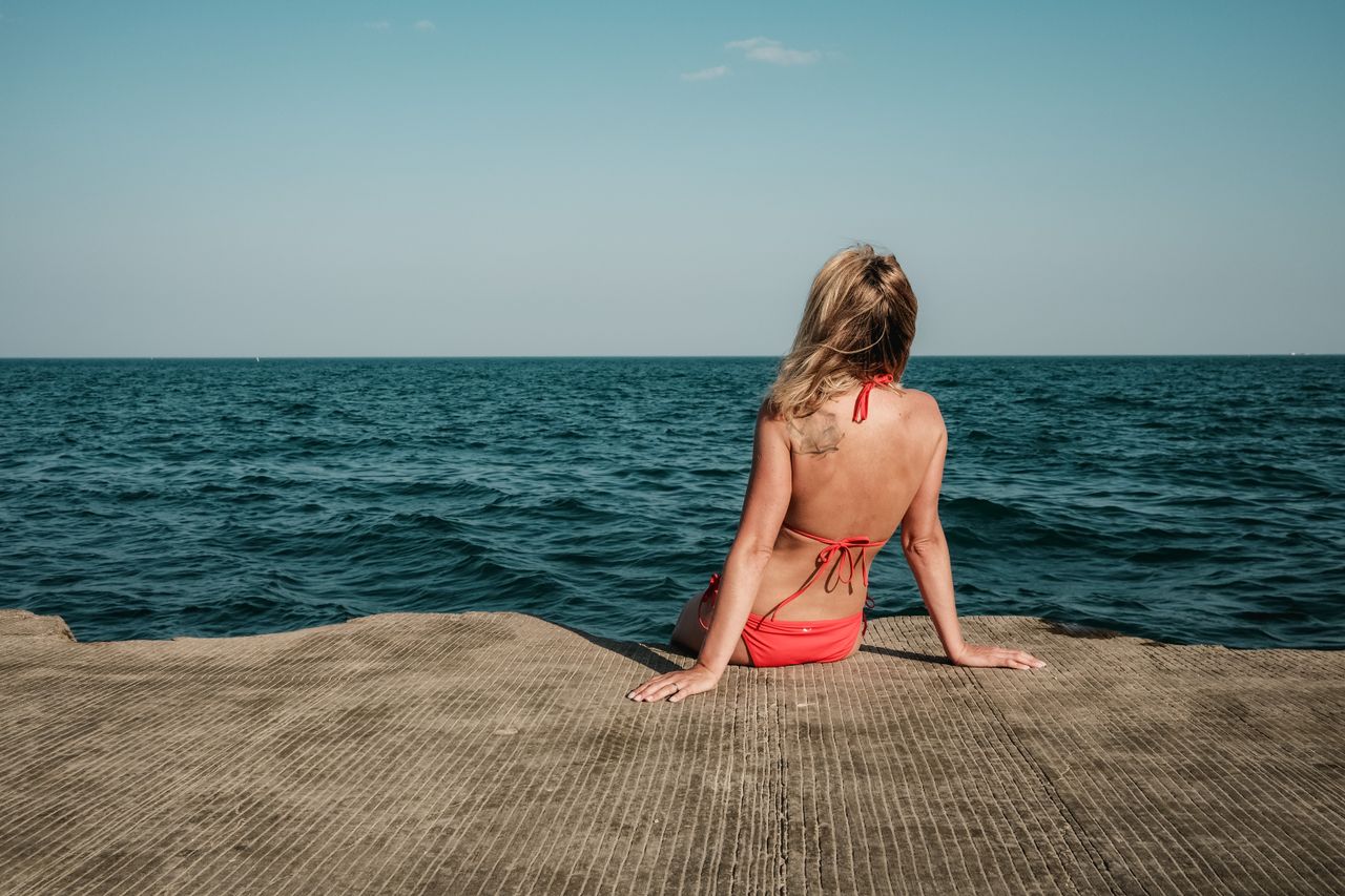 A woman in a red swimsuit sits on a pier overlooking the ocean, enjoying a sunny day.